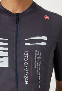 Cyklistický dres s krátkym rukávom v tmavošedej farbe s bielymi grafickými detailmi a červeným logom. Obsahuje predný zips a textilný materiál pre priedušnosť.