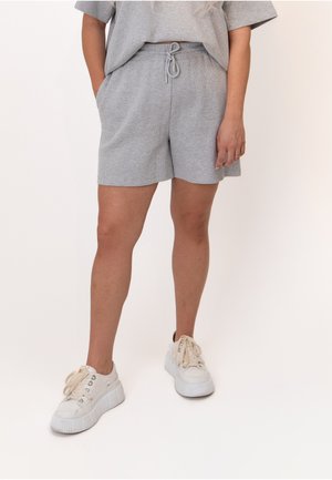Femme portant un short gris clair à cordon, un haut assorti et des baskets blanches à plateforme, debout avec les mains détendues.