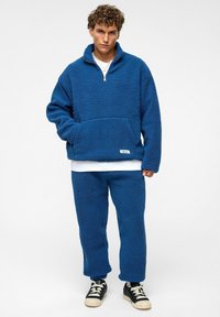 Blauwe fleece pullover met een halve rits, voorzak en een gestructureerd oppervlak, gecombineerd met bijpassende fleece broeken en zwarte sneakers.