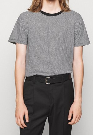 Personne aux cheveux longs portant une chemise à manches courtes noire et blanche rayée, rentrée dans un pantalon noir avec une ceinture noire.