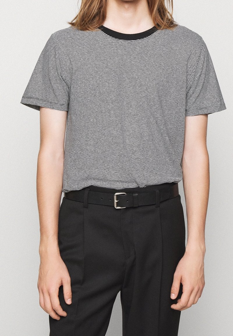 Personne aux cheveux longs portant une chemise à manches courtes noire et blanche rayée, rentrée dans un pantalon noir avec une ceinture noire.