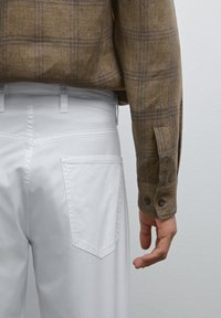 Bruine geruite overhemd met knoopmanchetten, gedragen met witte broek met een achterzak en stiksel. Hand rustend aan de zijkant.