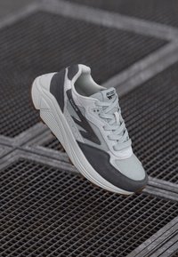 Chaussure de sport grise et blanche en matériaux mesh et daim, dotée d'une semelle épaisse, d'un laçage à l'avant et d'un logo discret sur la languette et le côté.