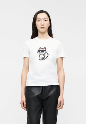 Ung kvinde iført hvid t-shirt med tegneserie-kat med solbriller på forsiden, kombineret med sorte læderbukser, stående mod en ensfarvet baggrund.