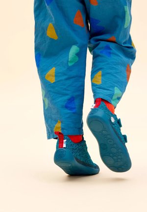 Scarpe per bambini color turchese con accenti rossi e strap in velcro. Indossate con pantaloni blu che presentano forme di cuori colorate e un tessuto testurizzato.