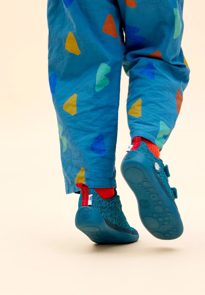 Scarpe per bambini color turchese con accenti rossi e strap in velcro. Indossate con pantaloni blu che presentano forme di cuori colorate e un tessuto testurizzato.