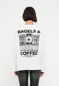 Άτομο που φοράει λευκή μακρυμάνικη μπλούζα με μαύρο σχέδιο χαρακτήρα μπέιγκελ σε πάγκο φούρνου και κείμενο «Μπέιγκελς & Καφές» στην πλάτη.
