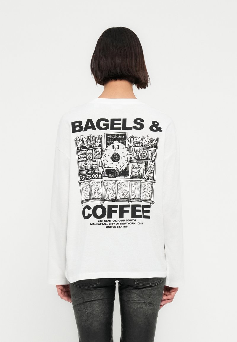 Άτομο που φοράει λευκή μακρυμάνικη μπλούζα με μαύρο σχέδιο χαρακτήρα μπέιγκελ σε πάγκο φούρνου και κείμενο «Μπέιγκελς & Καφές» στην πλάτη.