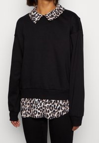 Mujer vistiendo un suéter negro sobre una camisa con cuello de estampado de leopardo, combinado con pantalones negros, de pie contra un fondo liso.