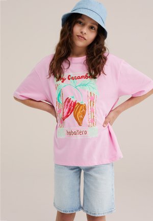 Młoda osoba nosząca różową, za dużą koszulkę z czerwonymi i pomarańczowymi paprykami oraz jasne szorty dżinsowe, ręce na biodrach i niebieski kapelusz typu bucket.