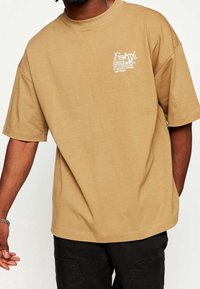 Camiseta oversized marrón de algodón, con un pequeño estampado gráfico blanco en el lado izquierdo del pecho y mangas cortas. Textura suave.