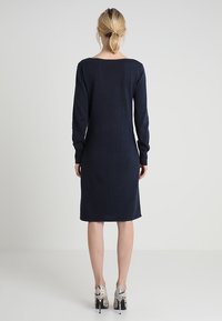 Navy blue long-sleeve dress, knee-length, soft fabric, slightly loose fit, with a wide neckline. Paired with patterned high-heeled shoes.