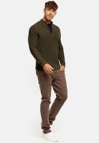 Cárdigan de punto texturizado en verde oliva con cremallera, combinado con pantalones ajustados marrones y zapatillas blancas. Esquema de colores cálido y neutro.