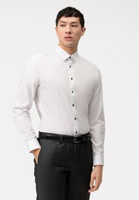 Camicia bianca a maniche lunghe con colletto classico, bottoni neri e texture liscia, abbinata a pantaloni scuri e una cintura.
