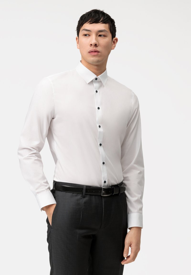 Camicia bianca a maniche lunghe con colletto classico, bottoni neri e texture liscia, abbinata a pantaloni scuri e una cintura.