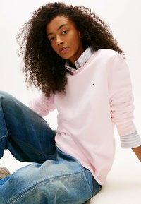 Felpa rosa chiaro con polsini a costine, indossata sopra una camicia a righe blu chiaro e bianca, abbinata a jeans denim larghi.
