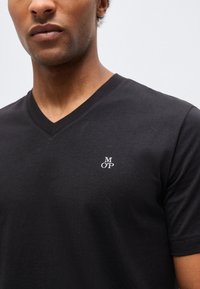 Άνδρας που φοράει μαύρο T-shirt με V λαιμόκοψη και μικρό λευκό λογότυπο "M O P" στο αριστερό στήθος, πάνω σε απλό ανοιχτό φόντο.