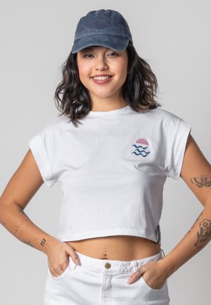 Lächelnde Frau in einer blauen Kappe, weißem Crop-Top mit Wellen- und Sonnenmotiv, weißen Hosen und sichtbaren Tattoos an beiden Armen und am Bauch.