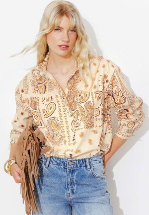Femme blonde portant une chemise beige à motif paisley rentrée dans un jean bleu taille haute, tenant un sac en daim marron à franges avec des bracelets au poignet.