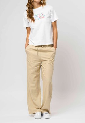 Ženska, oblečena v belo majico z rastlinskim potiskom, bež široke hlače z vezalko in belimi supergami, stoji z rokami v žepih na belem ozadju.