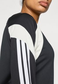 Juodas sportinis marškinėliai su baltomis ir pilkomis detalėmis; turi apvalią iškirptę ir tris vertikalias baltas juostas ant kairiojo rankovės. Apsiūtas audinys.