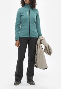 Tee-farbene Fleecejacke mit Reißverschluss und seitlichen Taschen; schwarze Hose und graue äußere Schicht. Getragen mit schwarzen Schuhen, die strukturierte Sohlen haben.