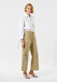 Beige weit geschnittene Hosen mit floraler Stickerei auf der linken Seite, kombiniert mit einem weißen Hemd mit Knopfleiste. Glatter Stoff und strukturierte Taschen.