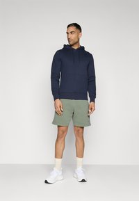 Sweatshirt azul-marinho com bolso frontal e cordões, combinado com calções verdes e ténis brancos com pormenores pretos.