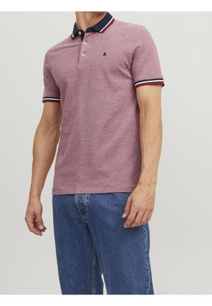 Mann trägt ein pinkfarbenes Polohemd mit kurzen Ärmeln, navyfarbenem Kragen und gestreiften Bündchen, kombiniert mit blauen Jeans, steht vor einfarbigem Hintergrund.