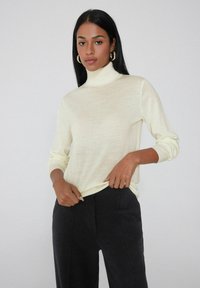 Suéter de cuello alto en crema, confeccionado en una tela suave, con mangas largas, dobladillo redondo y puños acanalados. Combinado con pantalones negros.