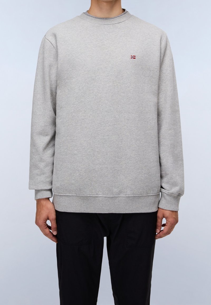 Homme portant un sweat-shirt gris clair à manches longues et col rond avec un petit emblème de drapeau rouge et blanc sur la poitrine, associé à un pantalon noir.
