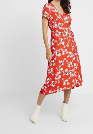 Femme portant une robe portefeuille rouge à fleurs avec des manches courtes et des bottines blanches, debout devant un fond clair uni.