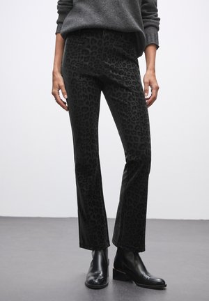 Schwarze Hose mit einem dezenten Leopardenmuster, aus einem weichen, strukturierten Stoff gefertigt. Ausgestellt geschnitten, kombiniert mit schwarzen Knöchelstiefeln.