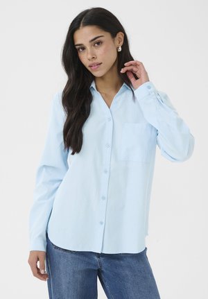 Mladá žena s dlhými tmavými vlasmi, oblečená v svetlomodrej košeli na gombíky a modrých džínsoch, pózujúca na jednoduchom bielom pozadí.