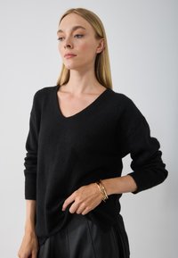 Pull en laine noir avec un col en V et des manches longues, présentant une coupe décontractée. Le mannequin porte un bracelet en or avec une jupe noire texturée.