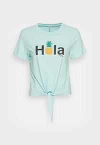 Světle modré tričko s krátkým rukávem a uvázaným předním dílem s textem "Hola", kde je písmeno "o" nahrazeno grafikou ananasu a je zde také malé slovo "tropical".
