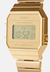 Orologio digitale dorato con quadrante quadrato, display LCD che mostra l'ora e la data, e un bracciale in metallo. Caratteristiche di resistenza all'acqua e funzione sveglia.