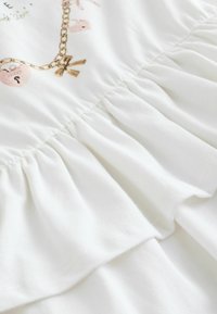 Biela látka s volánovým dizajnom, zdobená vzorom zlatých retiazok s príveskami ako mašľa a zámok. Hladká textúra s jemným leskom.