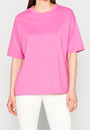 Kobieta w gładkim, jasnoróżowym, oversize T-shircie z krótkim rękawem i białych spodniach, stojąca na jasnym tle.