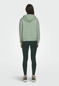 Žena stojící zády k fotoaparátu, oblečená v světle zelené mikině, tmavě zelených legínách a zeleno-bílých teniskách na jednobarevném pozadí.