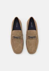 Tan suede slip-on shoes featuring a decorative metal chain accent across the upper and a smooth texture with a rounded toe design.