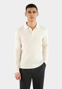 Helle beige, gerippte Langarm-Polohemd mit Kragen, in einer taillierten Passform und subtiler Struktur. Kombiniert mit dunkel gestreiften Hosen.