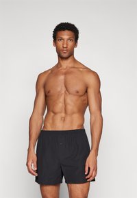 Black cotton boxer shorts with an elastic waistband and a single button fly, worn by a shirtless man with a muscular build.