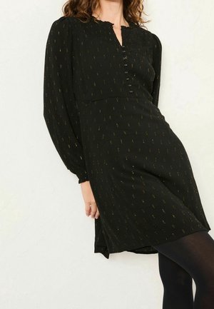 Femme portant une robe noire à manches longues jusqu'aux genoux avec petits motifs dorés et boutons sur le devant, associée à des collants noirs.