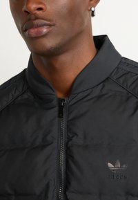 Schwarze gepolsterte Jacke mit Stehkragen, Reißverschluss und dezentem schwarzen Adidas-Logo auf der linken Brust. Glatte Stoffoberfläche.