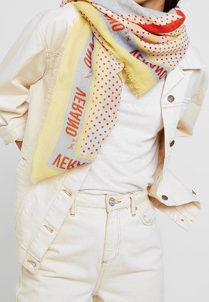 Femme portant une veste et un pantalon en denim crème avec une chemise blanche, accessoirisée d'une écharpe multicolore avec texte rouge et pois.