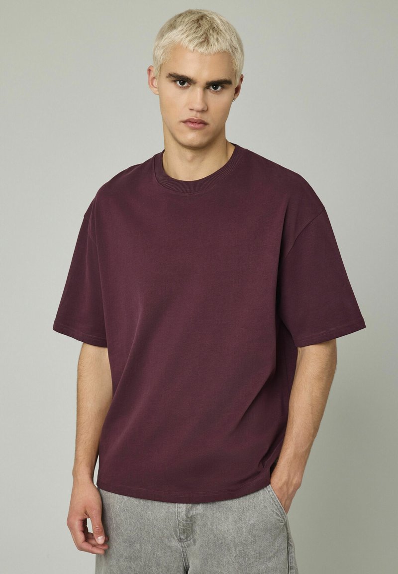 T-shirt oversize bordeaux en coton avec un col rond et des manches courtes. Texture lisse, design minimaliste et coupe décontractée.