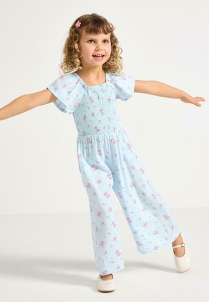 Ung pige balancerer med arme udstrakt, iført lyseblå blomsterbuket jumpsuit og hvide sko på ensfarvet baggrund.