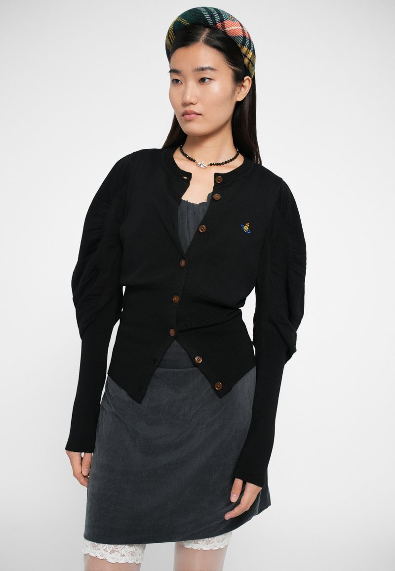 Cardigan nero con maniche a sbuffo, bottoni in legno e dettagli ricamati blu, abbinato a una gonna grigia con accenti in pizzo.