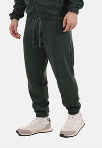 Pantaloni jogger in velluto a coste di colore verde scuro, con vita e polsini elastici; abbinati a sneakers chiare con una palette di colori neutra e accenti testurizzati.